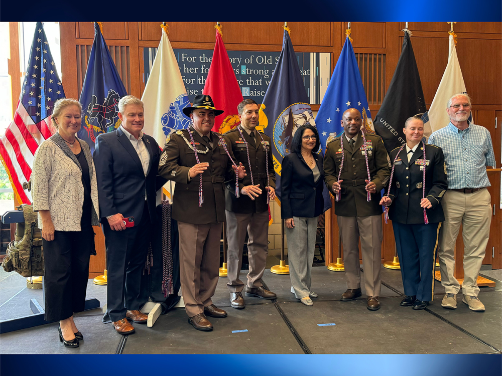 A group of people pose in front of the American flag and U.S. military flags.