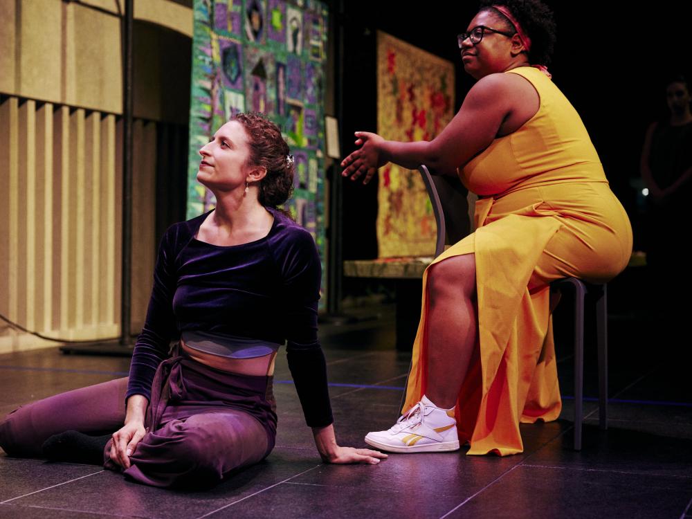 A white woman sits straight on a floor stage next to a Black woman sitting in a chair.