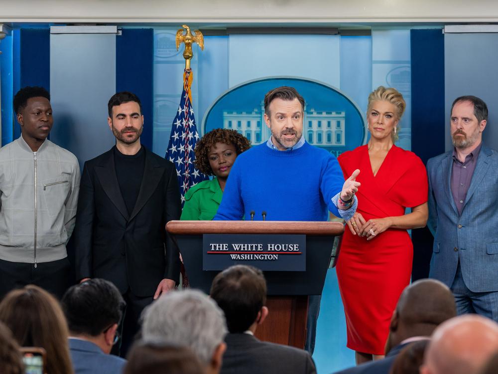Six people stand behind a podium bearing a sign that reads "The White House"