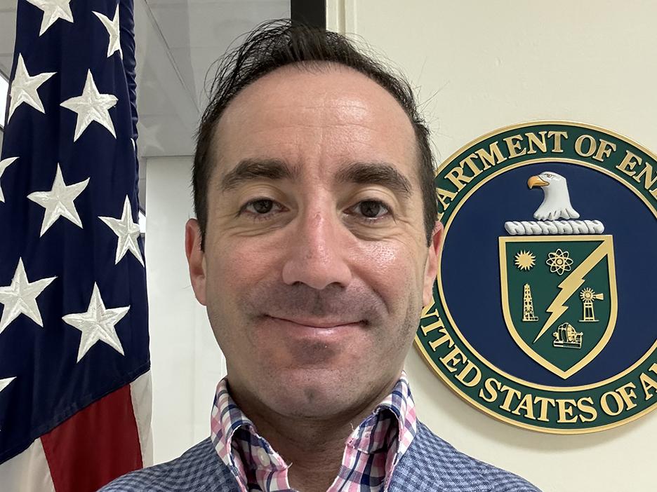 A man standing between a U.S. flag and a Department of Energy seal.