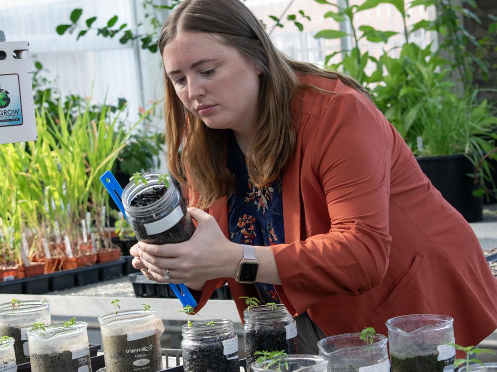 A woman holds a jar of soil and a ruler, taking a measurement, with plants behind her