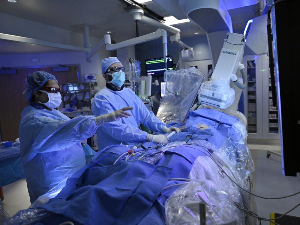 Two physicians perform a procedure on a draped patient in a surgical setting. Various pieces of medical equipment are located throughout the room.