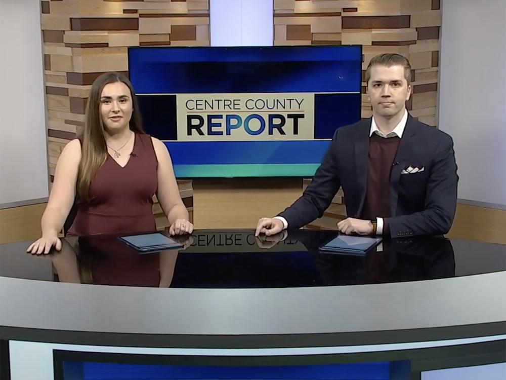 A woman in plum dress and a man in a coat and sweater vest sit behind a TV news desk