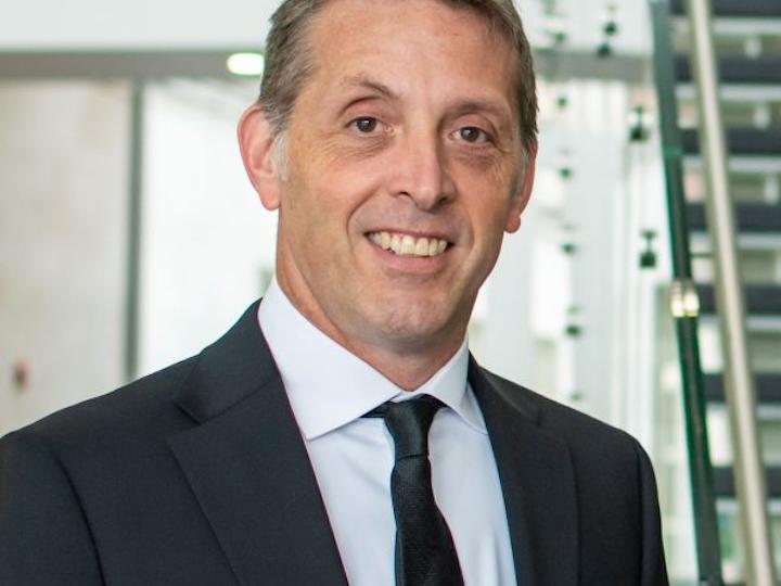 Christian Connell headshot with black jacket, black tie, white shirt, and short graying brown hair, smiling.