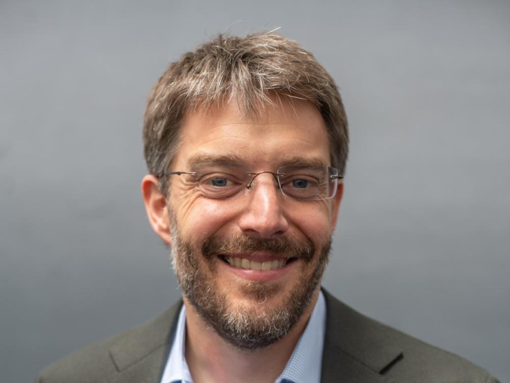 Dean Spears headshot in brown suit jacket, light blue button-down shirt, brown hair and beard, glasses, smiling.