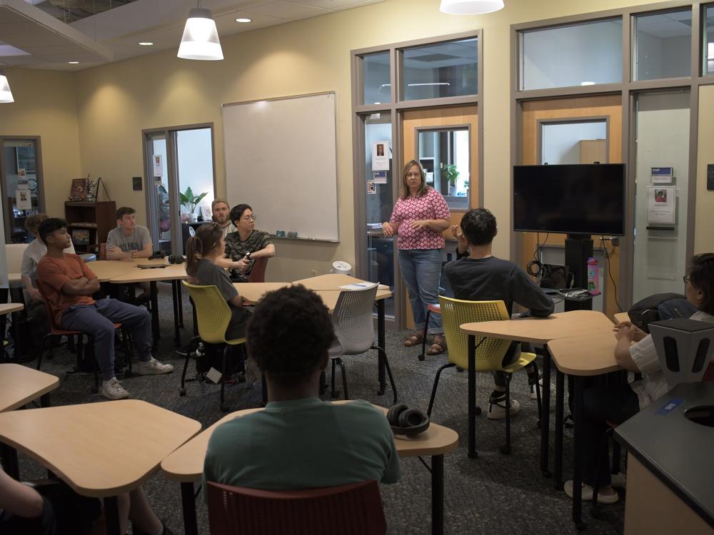 Person stands at the front of a room, talking to other people sitting at tables. 