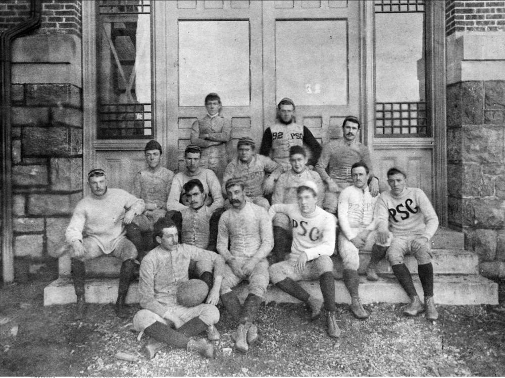 A photo of the football team, with most being seated on steps. The two in the back are leaning against a door.