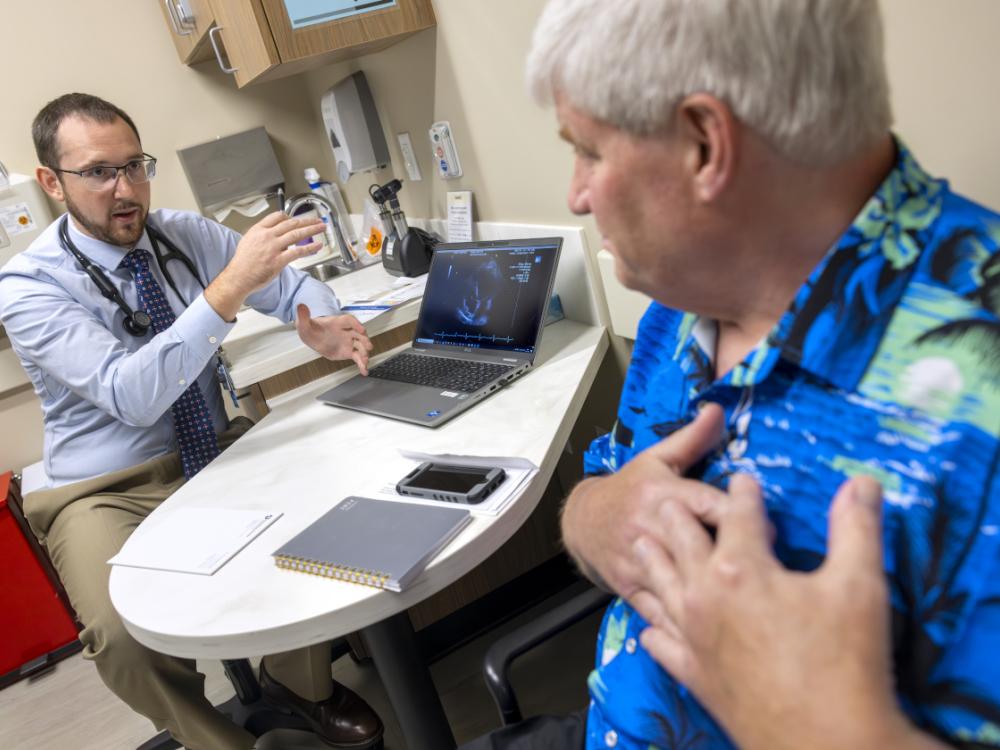 Tyler Thomas, left, gestures as he talks to patient John Jones, right. Thomas is wearing a button-down shirt, tie, glasses and has a stethoscope around his neck. Jones is wearing a Hawaiian shirt and has hands over his heart.