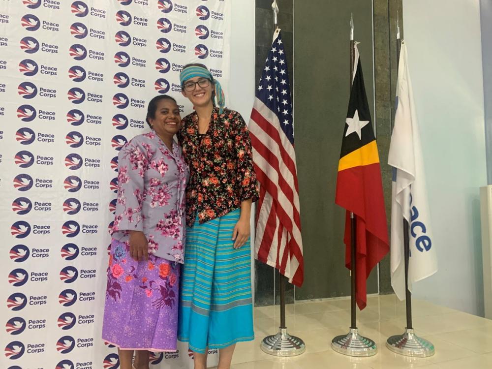 Two people smile, arm-in-arm, wearing colorful garb in front of a Peace Corps banner