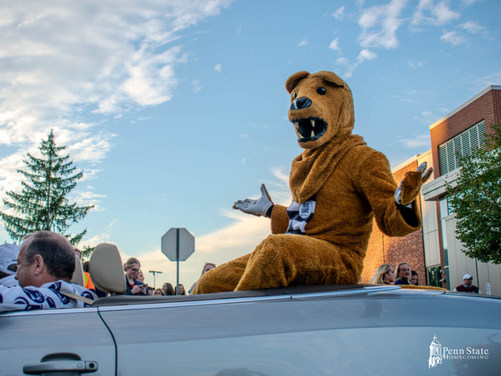 The Nittany Lion mascot sits in the back of a convertible, with a partly cloudy sky and a brick building in the background.