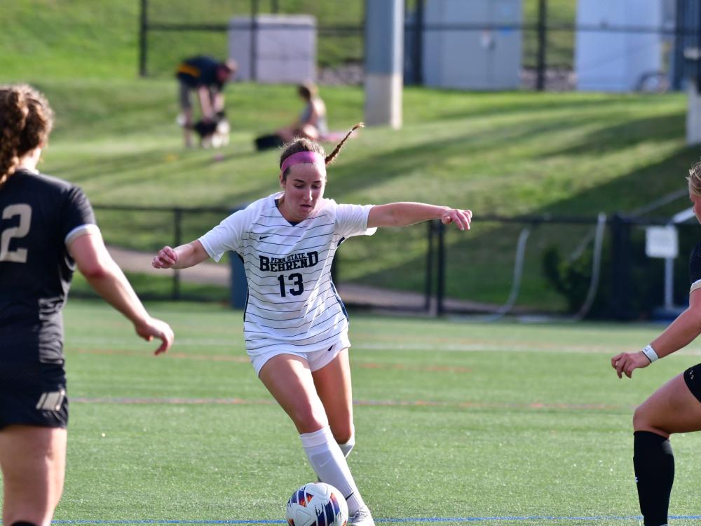 A Penn State Behrend soccer player dribbles between two defenders.