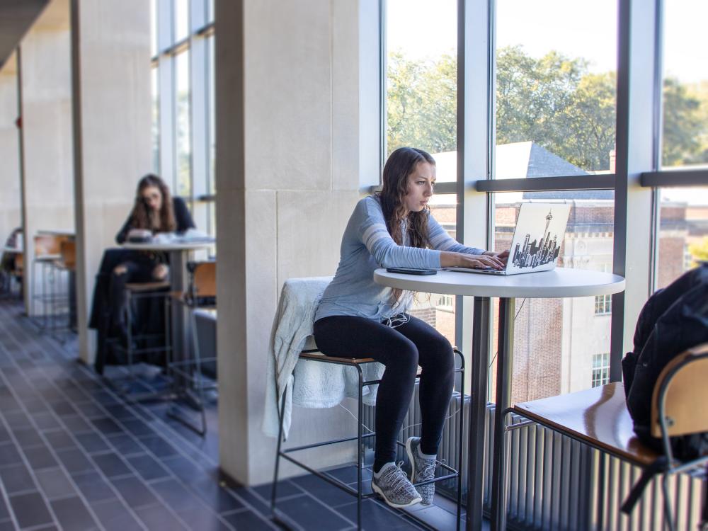 A student sitting at a high-top study table with a laptop against a wall of windows looking out over campus.