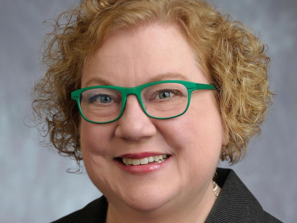 Head shot of a woman with blonde curly hair and green-rimmed glasses smilng