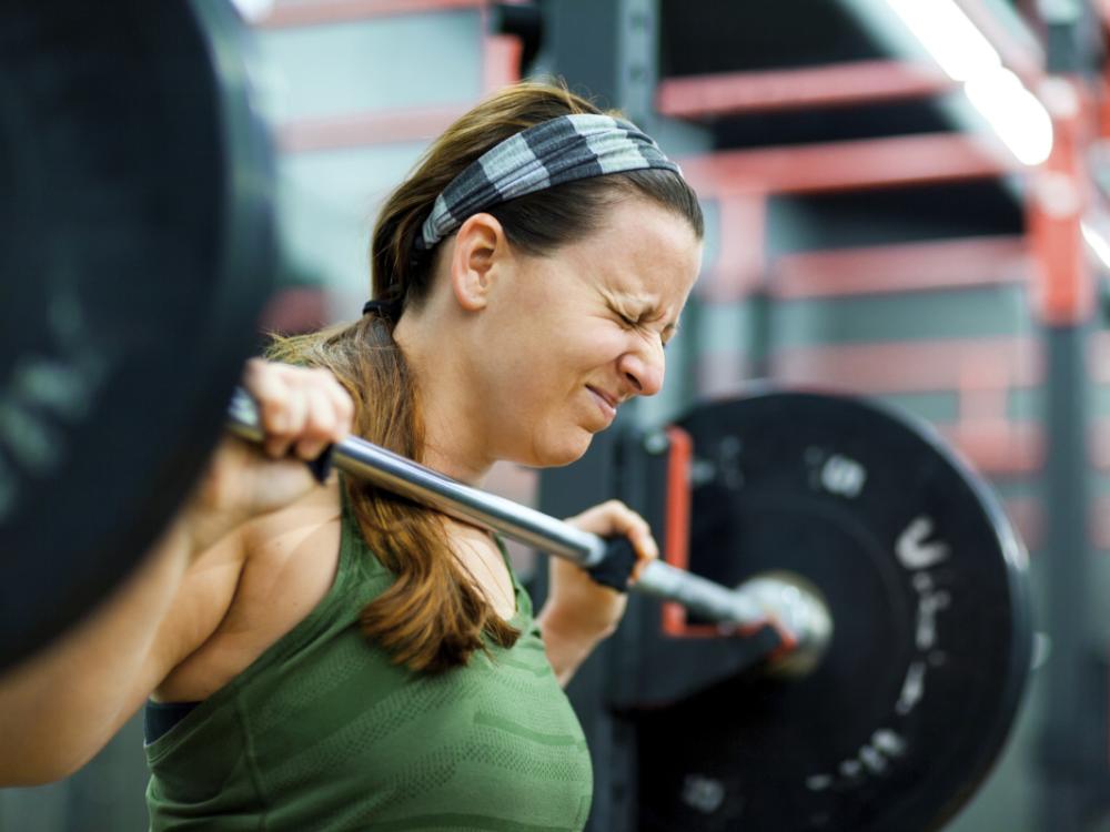 A woman closes her eyes as she lifts a heavy weight off of a rack at a gym.