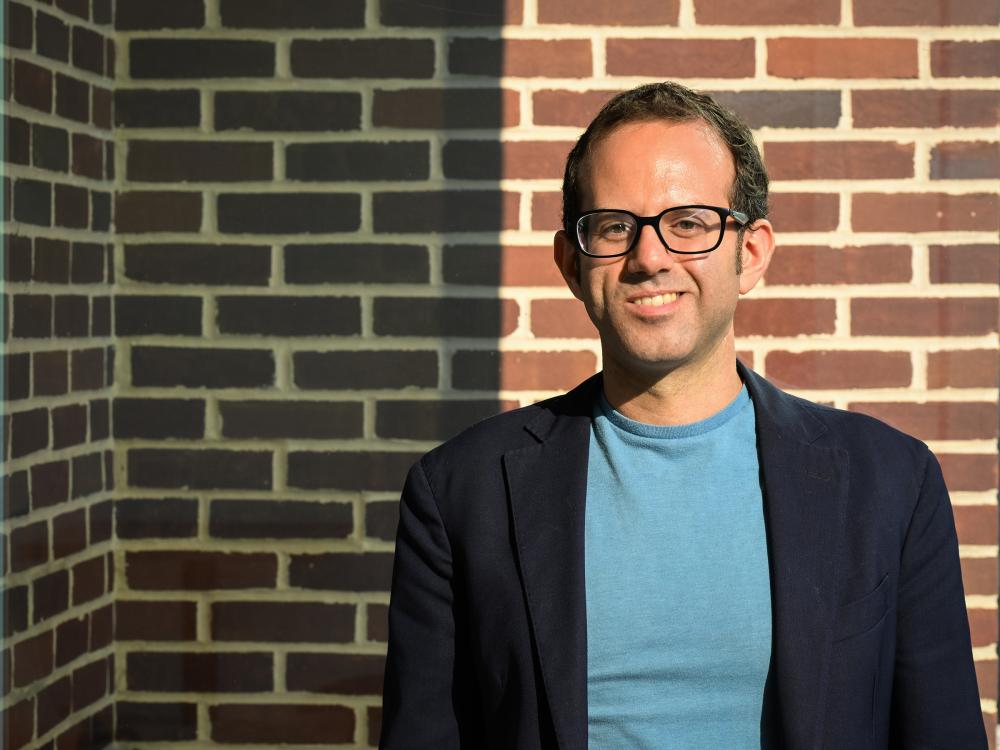 A person wearing glasses stands in front of a brick wall outside and smiles at the camera 