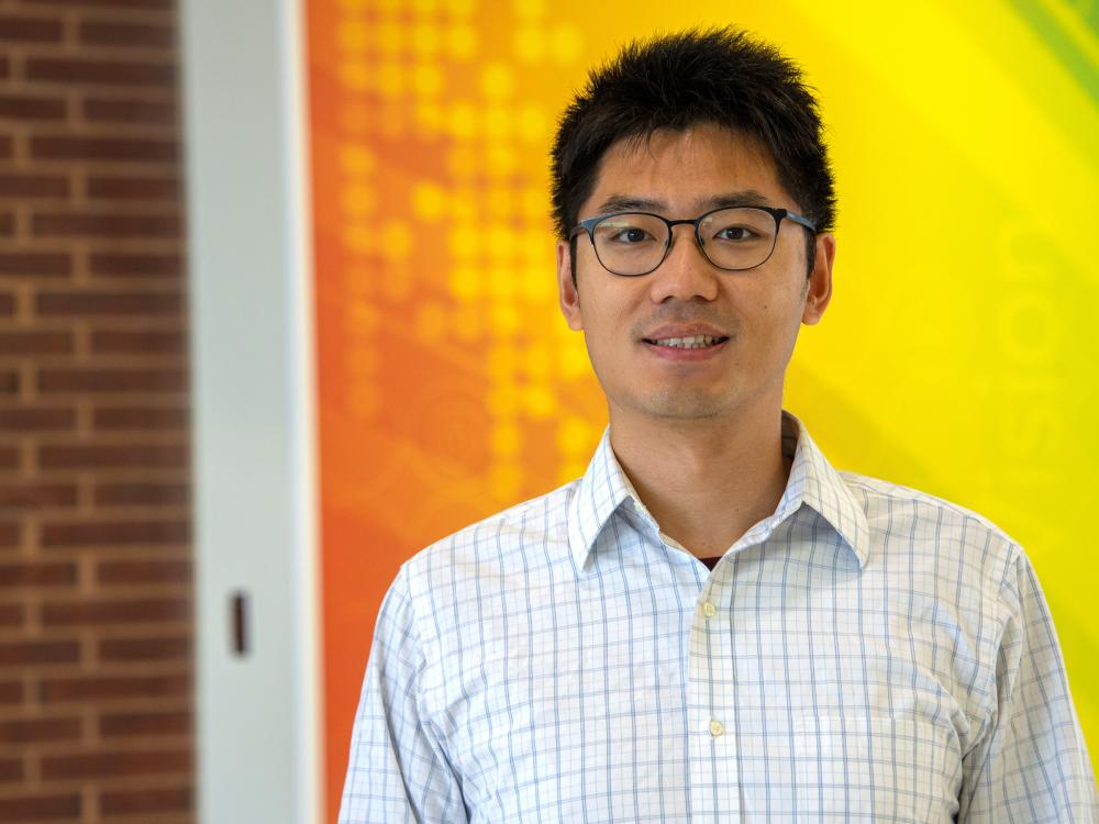 A portrait of an individual standing behind a brick, orange, green and yellow backdrop. 