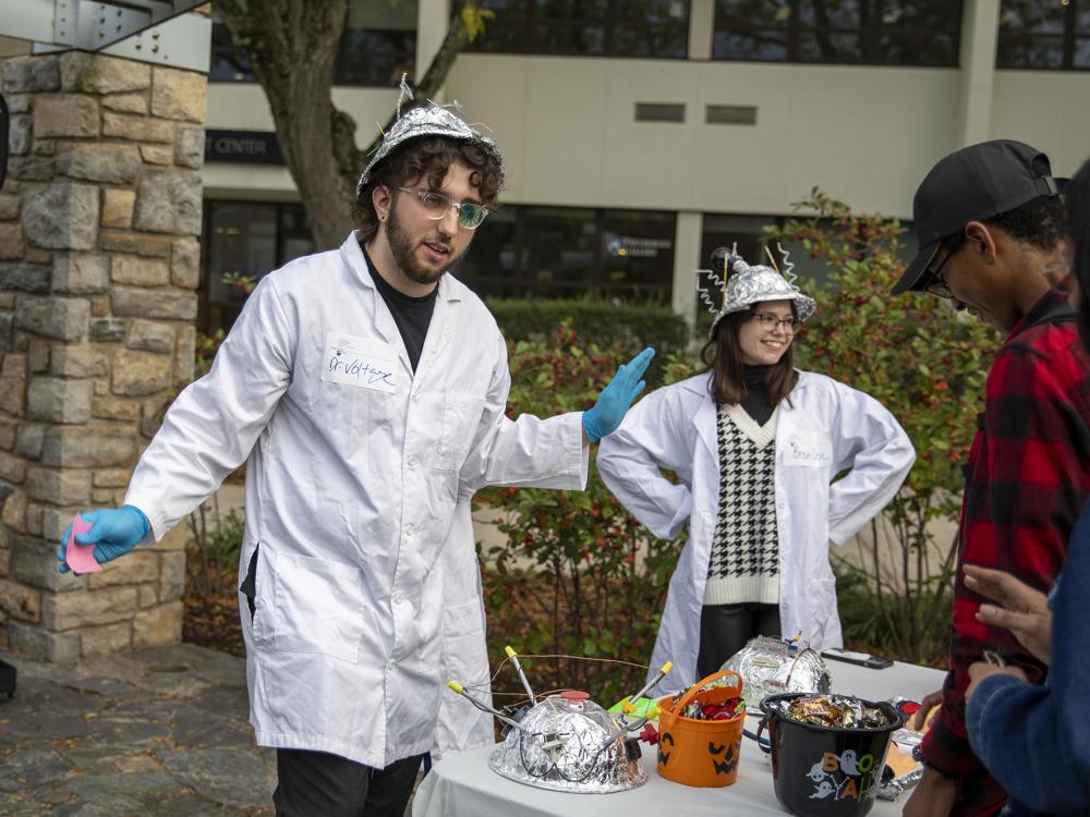 Students wearing lab coats and tinfoil hats conduct experiments with onlookers