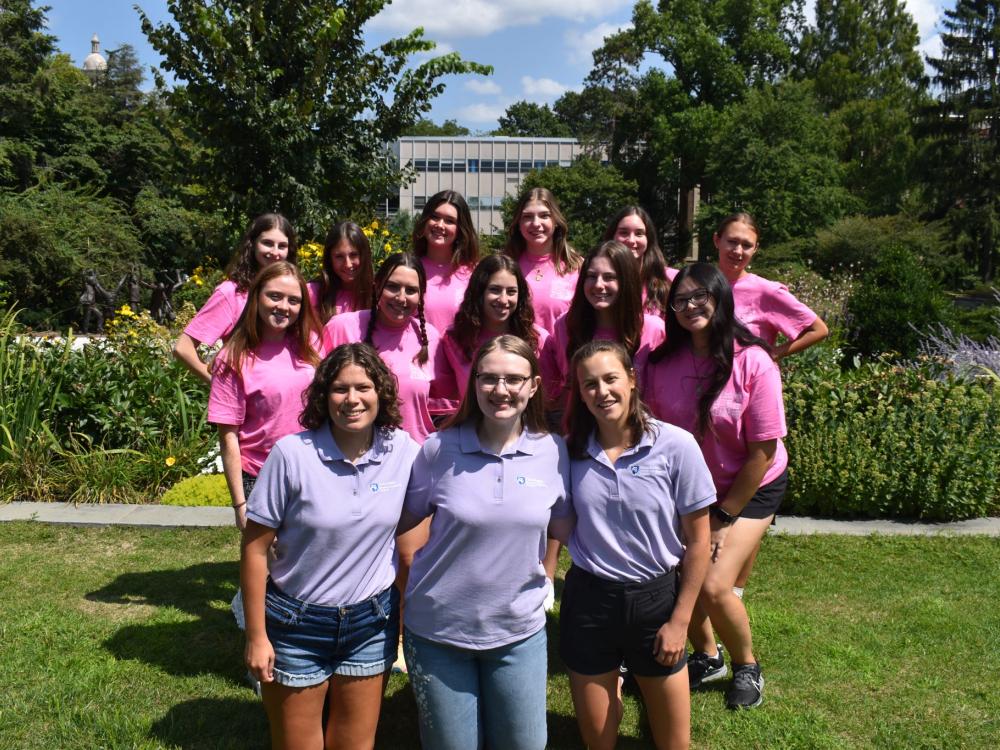 Women wearing pink and purple tee shirts standing together in the sun, in front of trees, posing for a photo