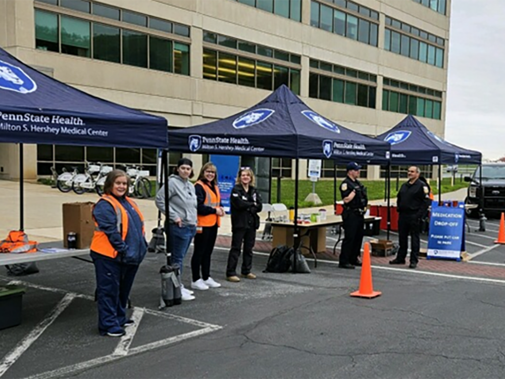 Six people stand near canopies with Penn State Health logos on them. A large building is in the background.