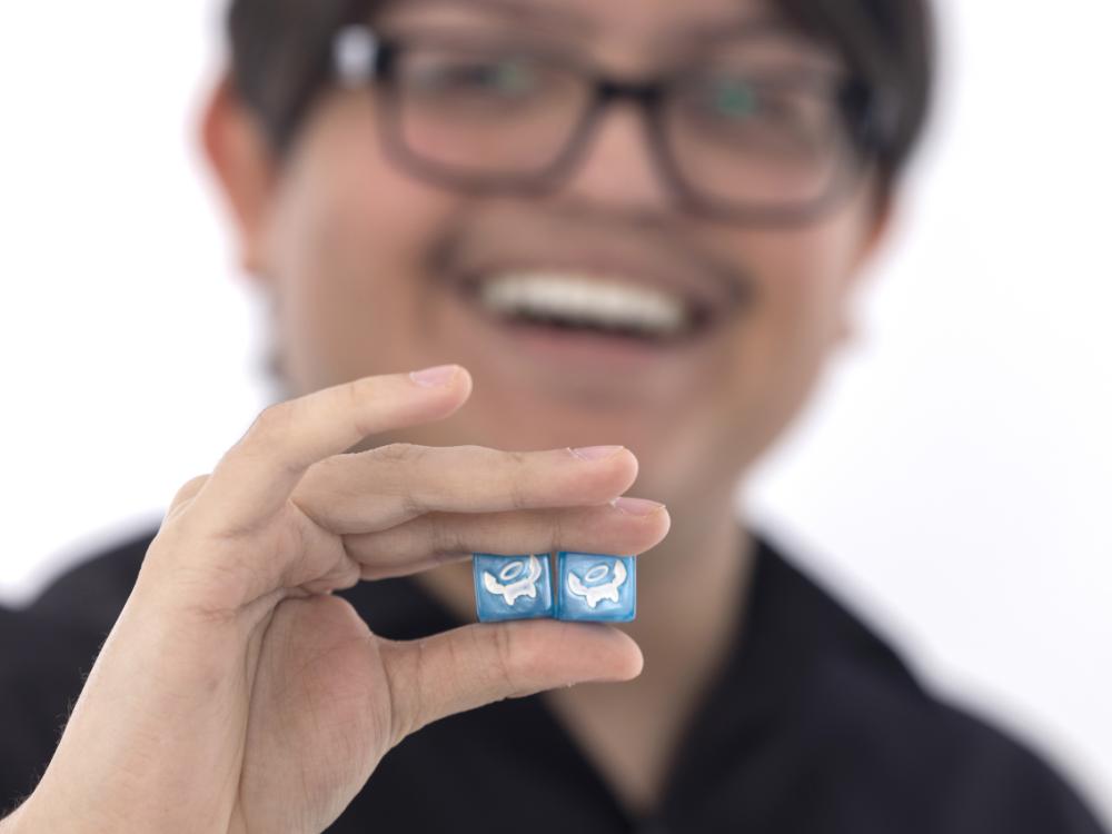 Joseph Romero poses with a pair of specially designed dice that he’s holding near the camera. Joseph is in the near background, slightly out of focus, smiling.