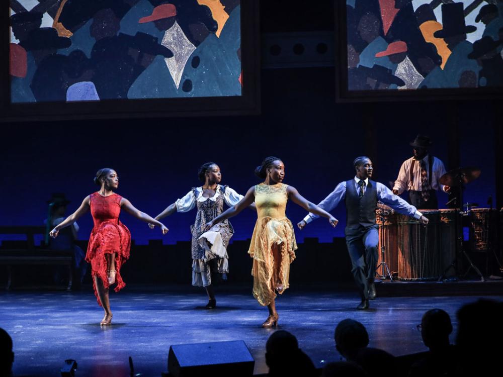 Black dancers strike matching poses while a backdrop shows bold paintings.