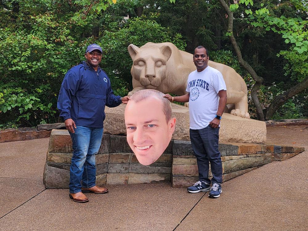 Two men pose in front of the Nittany Lion Shrine with a printed cutout of a man's head.