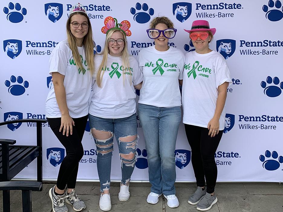 A group of four women, one in oversized glasses, stand against a backdrop with the Penn State Wilkes-Barre logo.