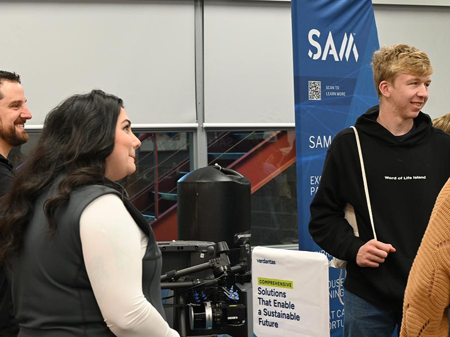 Adults and students conversing during a career fair, with a flag with the SAM logo at back right.