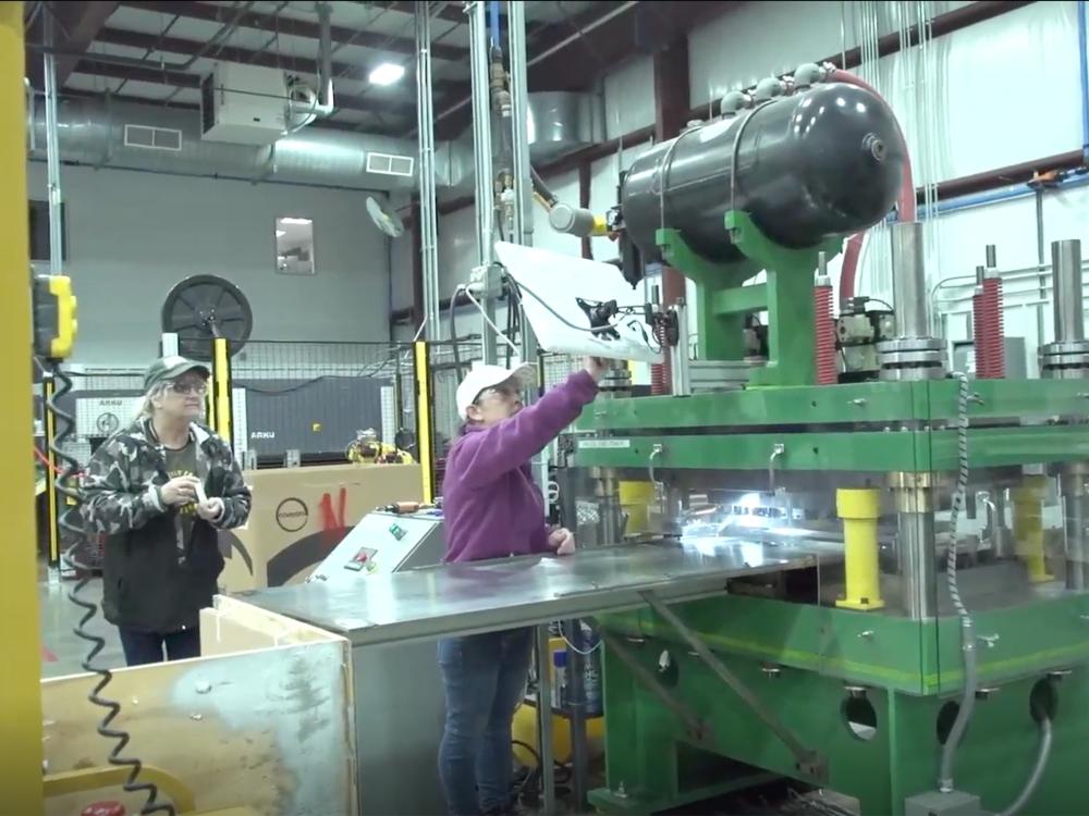 Two people in baseball caps and safety glasses work in a manufacturing plant 