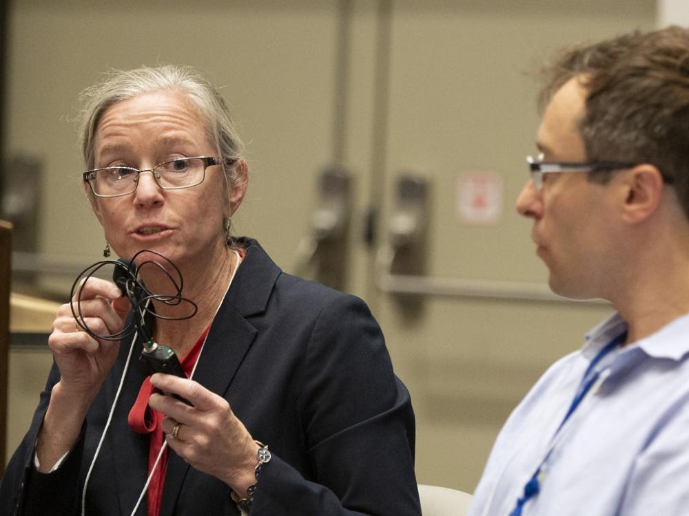 A woman holds a microphone next to her face while she speaks. She is sitting next to a man.