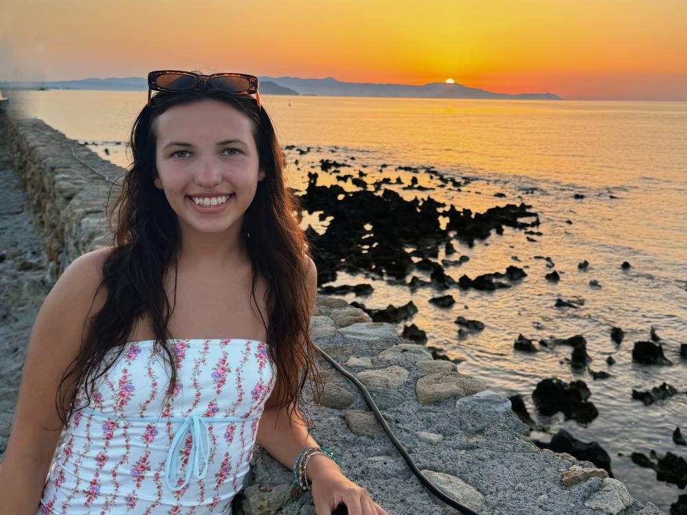 Elisabeth Egeli stands next to the ocean overlooking a sunset in Greece.