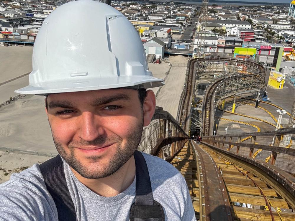 Eli Rush wearing a white hard hat while climbing a roller coaster hill. A town is in the background.