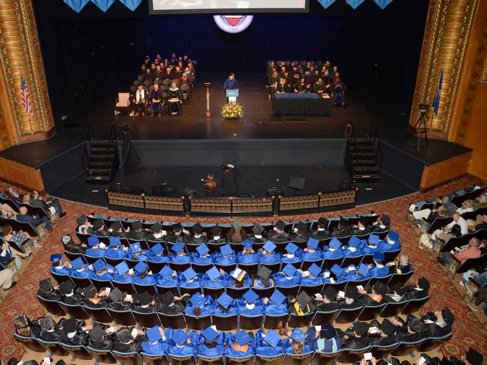 A balcony view of a theater stage and seats filled with people in blue graduation caps and gowns