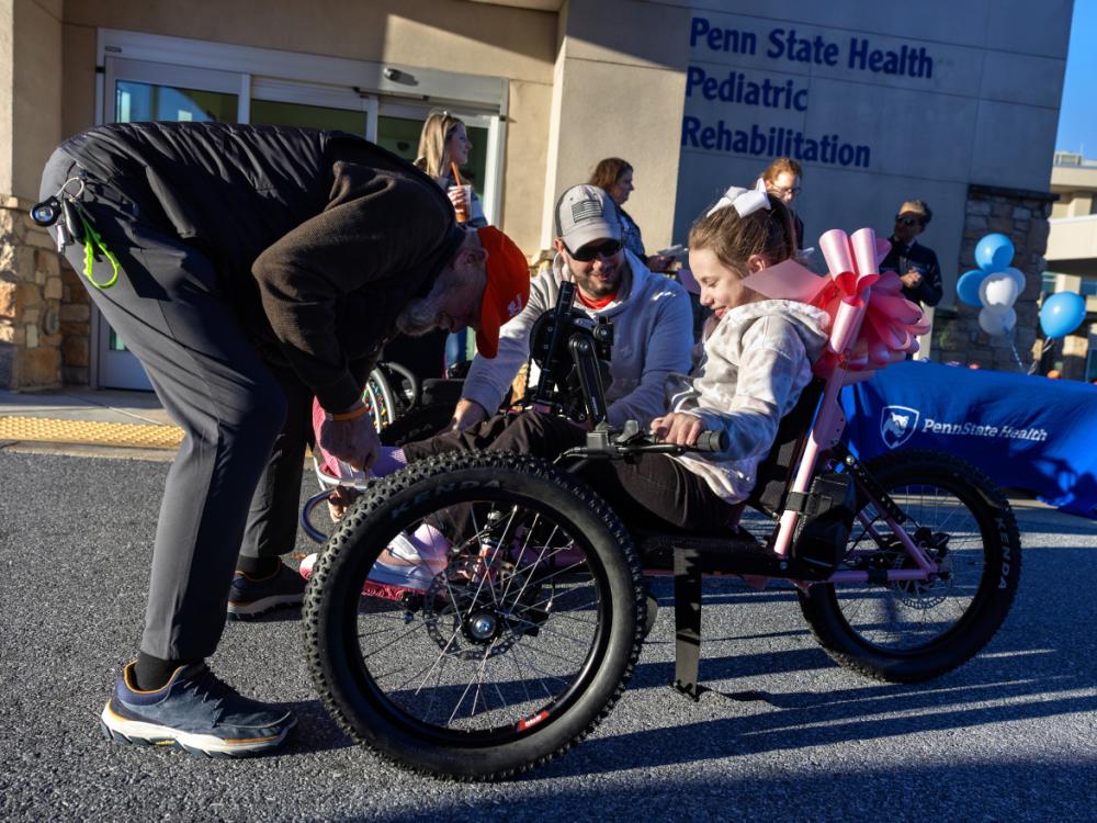 A man leans over to adjust the front of an adaptive bicycle. A young girl sits on the bike smiling, as her dad looks on. Additional people stand in the background.