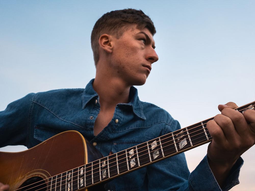 A young white man holds an acoustic guitar and looks off into the distance.