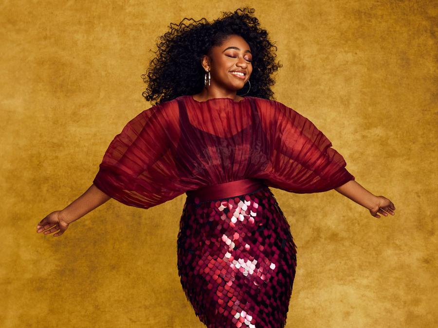 A young Black woman wearing a sequinned dress stretches out her arms.