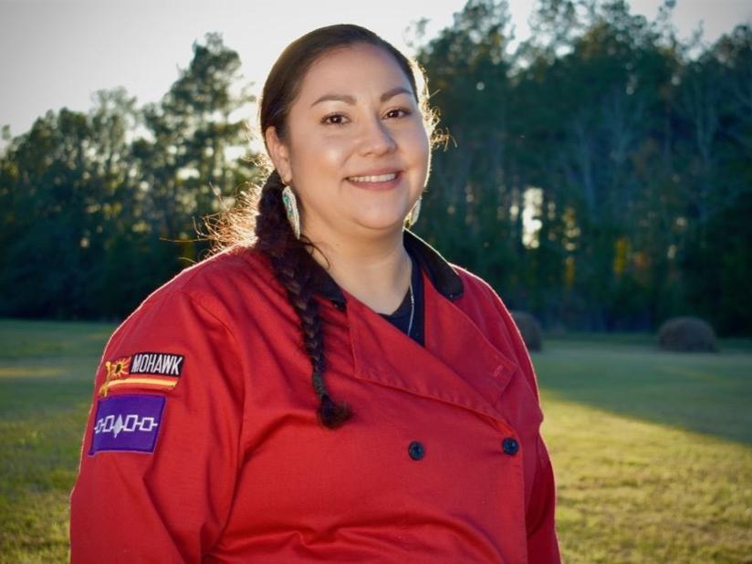 Tawnya Brant stands in a field wearing a long braid and a red Chef jacket.