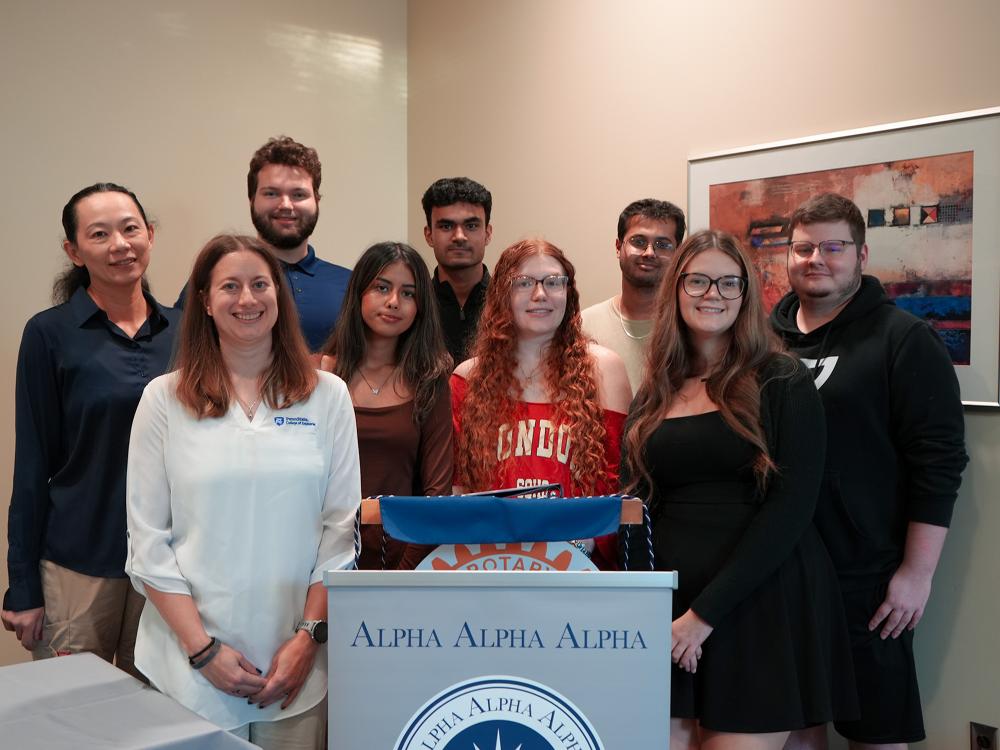 Smiling students and faculty members stand together behind an Alpha Alpha Alpha (Tri-Alpha) Honor Society podium