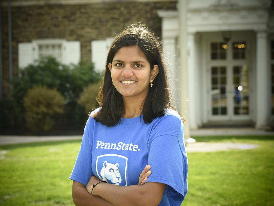 A student wearing a blue shirt with the Penn State mark, standing in front of a historic building.