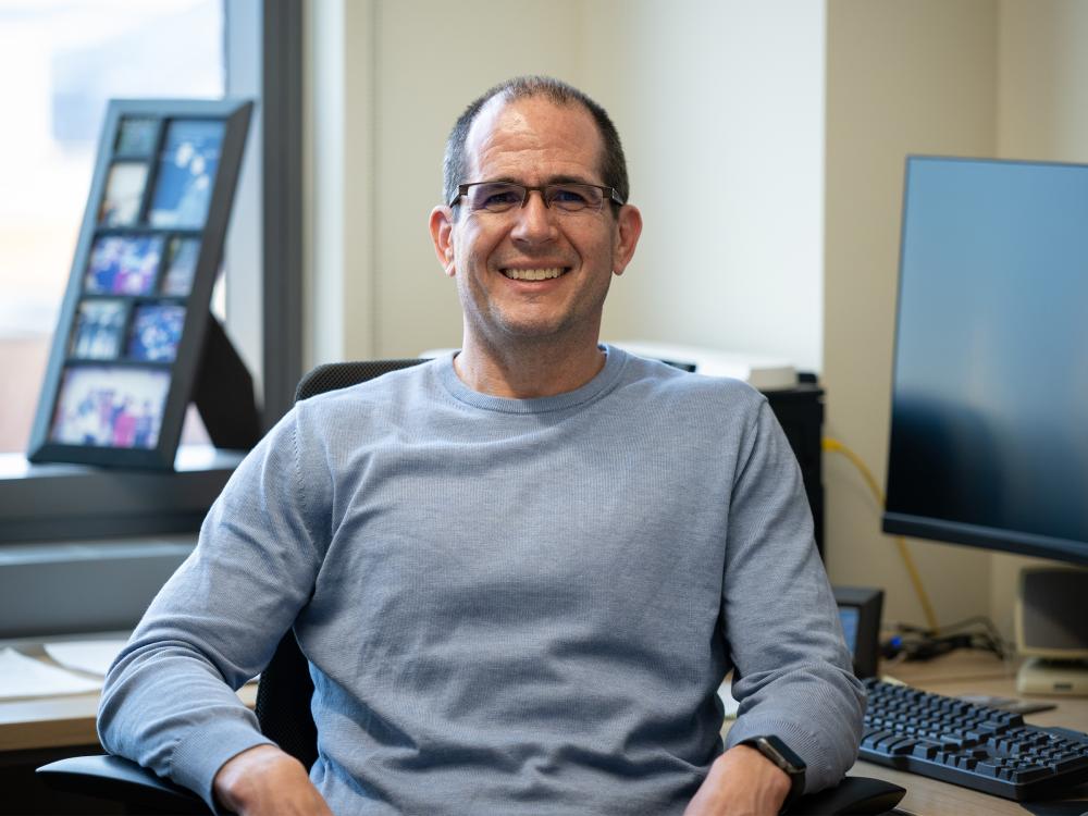 A person in glasses sits at a desk with a computer monitor in the background 