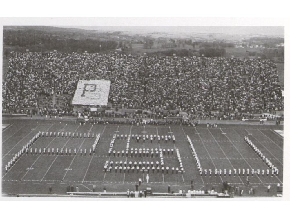 A marching band forms "PSU" on a football field in front of a crowd