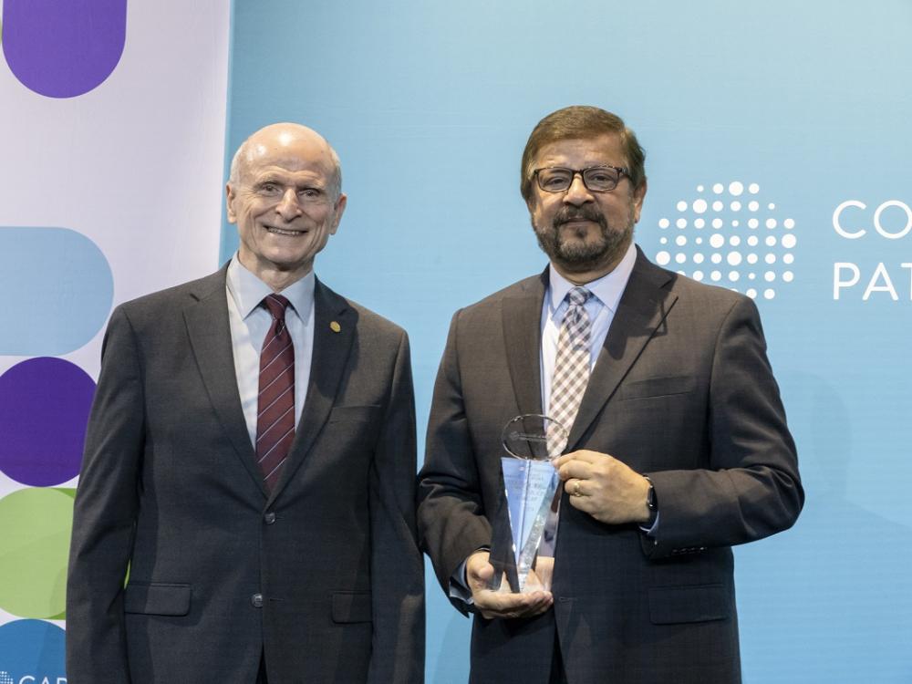 Two men stand on a stage posing for a photo. The man on the right is holding an award.