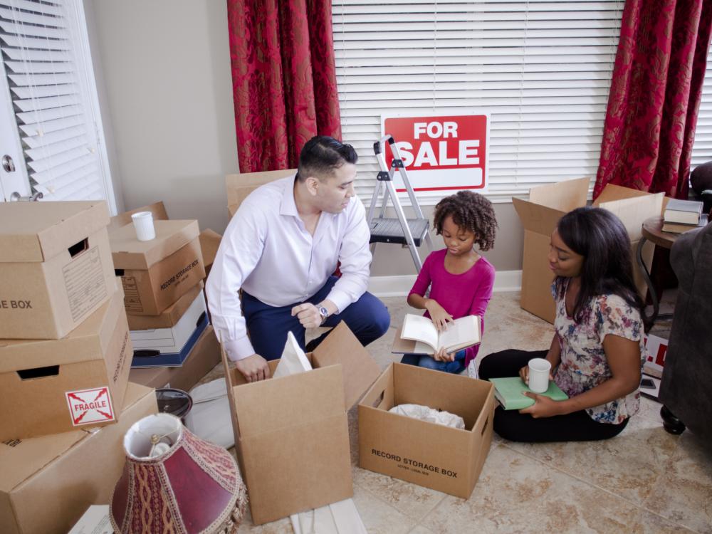 Multicultural family members pack boxes in new home. “For Sale” sign hangs on wall..