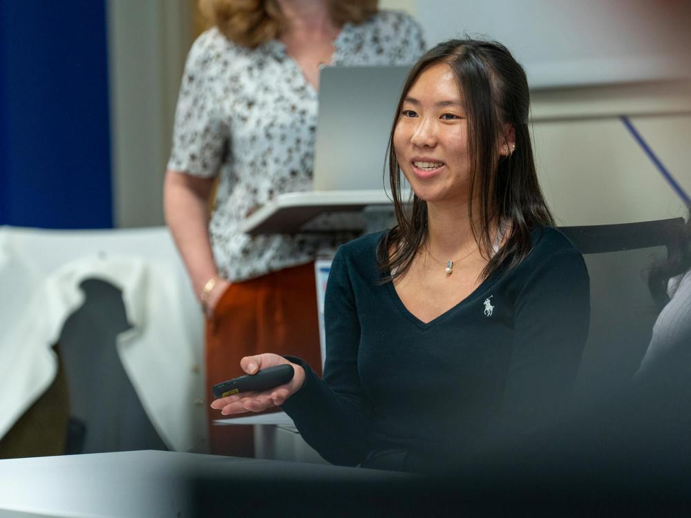 Charis Liu presents while sitting behind a desk at the front of a classroom.