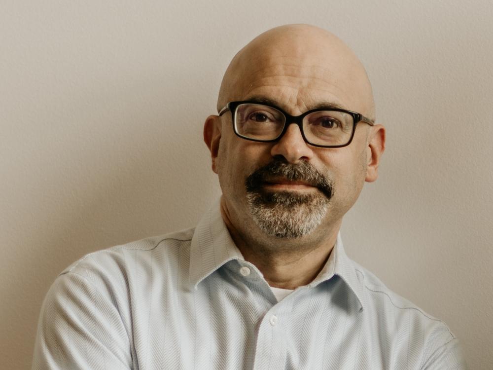 Image of a white man with a bald head wearing black framed glasses with his arms casually crossed.