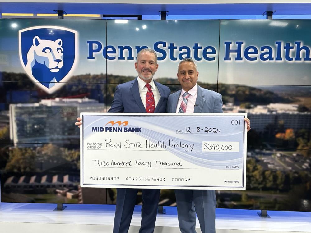 Two men in suits pose with a check made out to “Penn State Health Urology” in the amount of $340,000. A large Penn State Health logo is in the background.