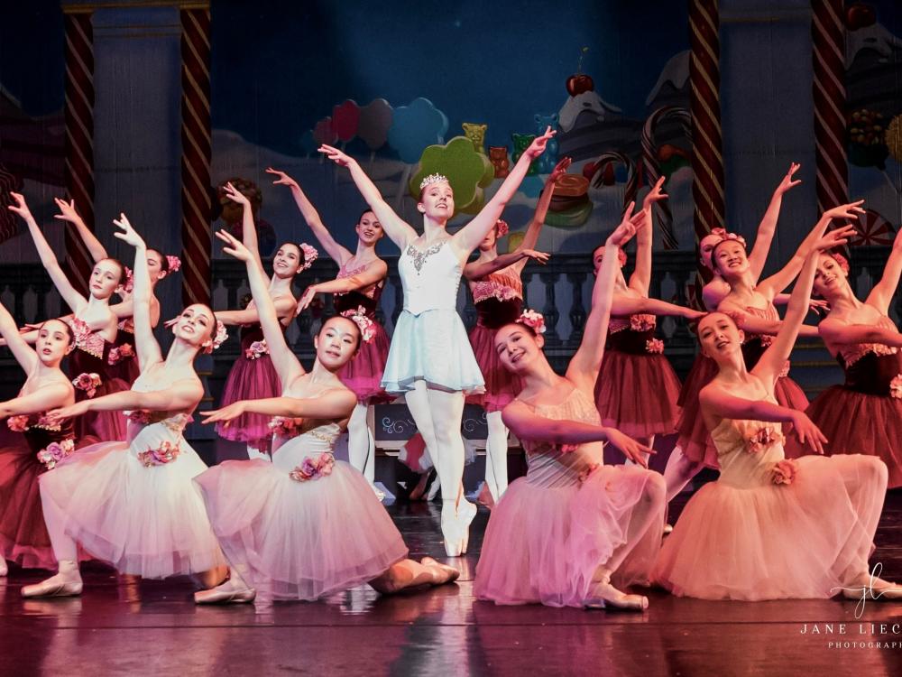 A group of young ballet dancers wear matching tutus and crowd around a dancer in the center in the pointe position.