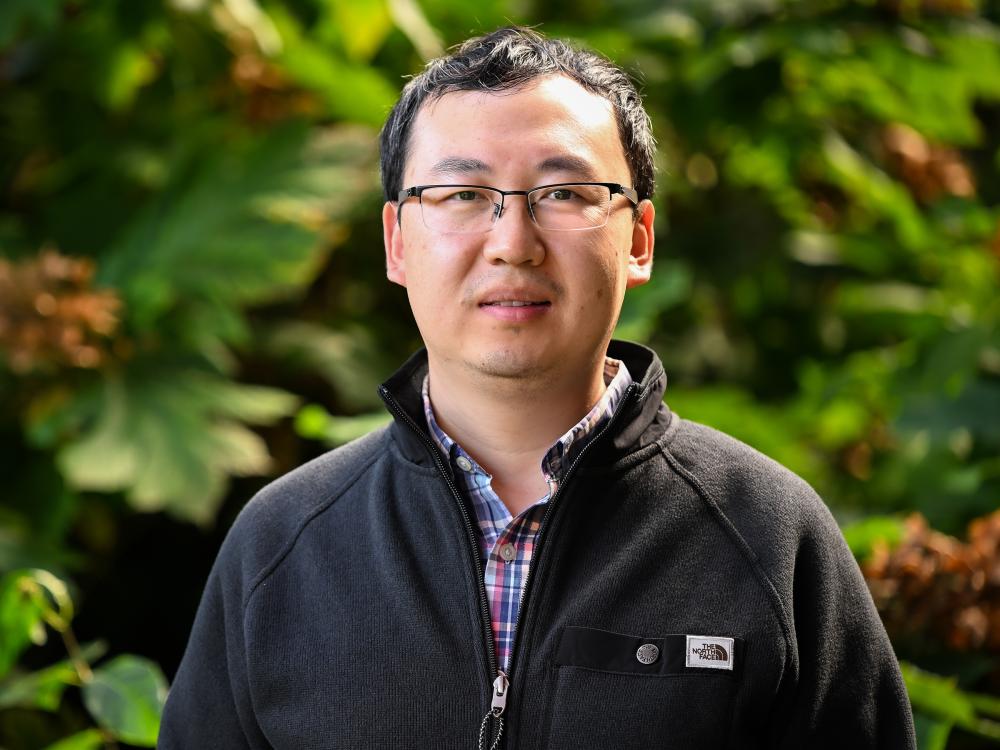 An individual poses outdoors in front of trees.