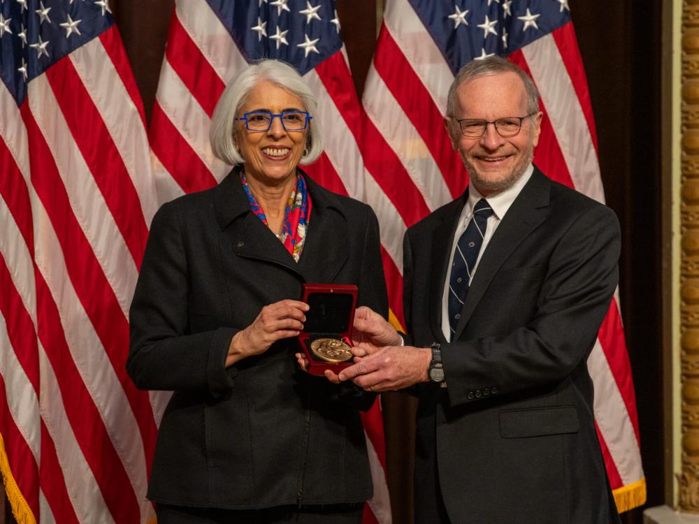 Two people stand in front of the American flag holding a medal