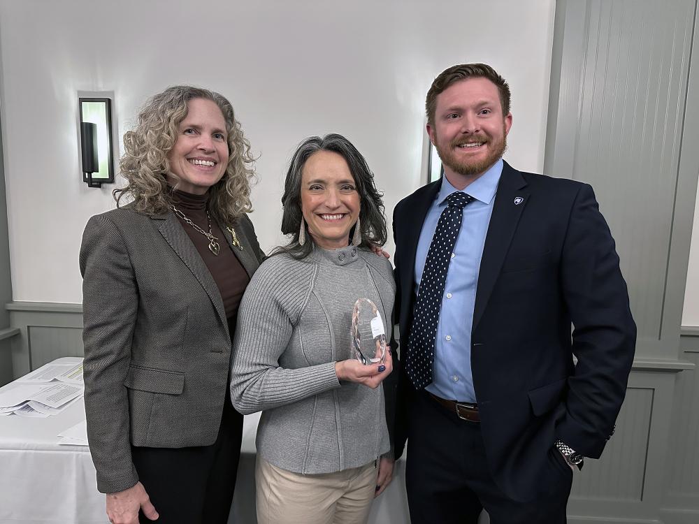 A photo of three people, and the woman in the center is holding a crystal award